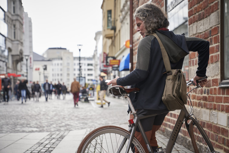 Langhåret mann i turklær, med sekk over ryggen, lener seg til sykkelen sin i København mens han surfer på mobilen sin
