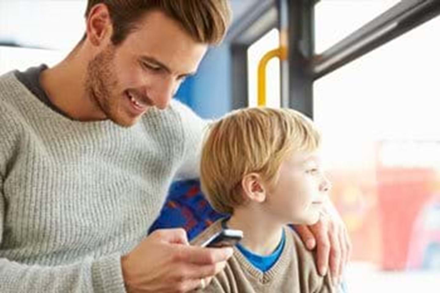 En foresatt og et barn sitter på bussen. Faren ser på mobilen hans, der han kommuniserer med læreren på skolen via skole-appen.