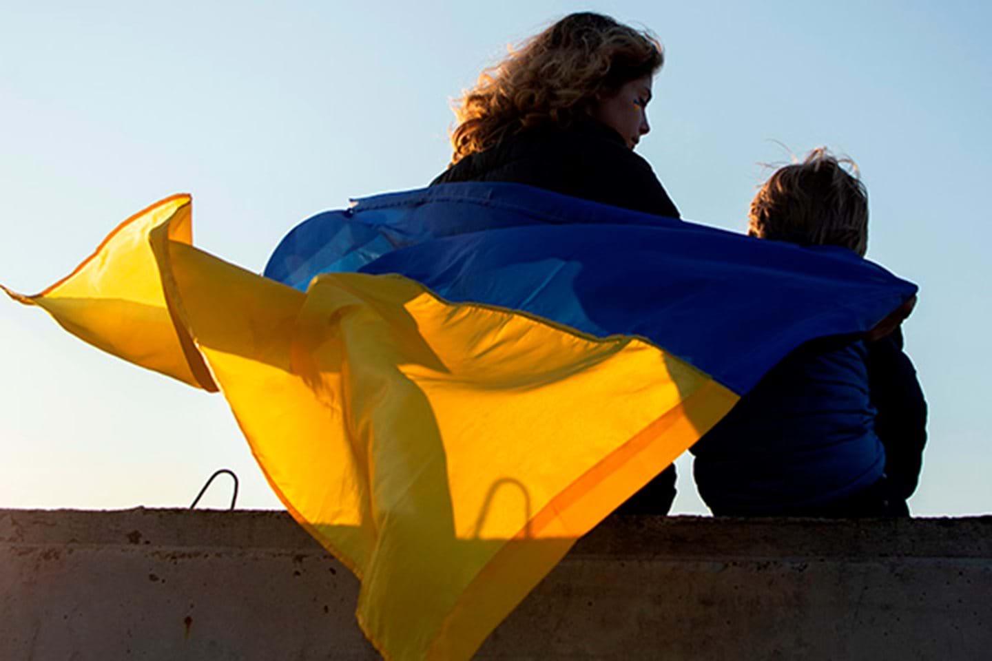 To personer sitter på en betongmur med et stort Ukrainsk flagg over seg mot en blå himmel. Bildet representerer bosetting av Ukrainske flyktninger.