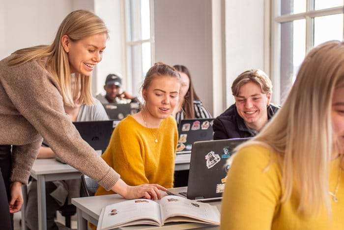Kvinnelig lærer i et videregående klasserom som hjelper elever som jobber på laptop.